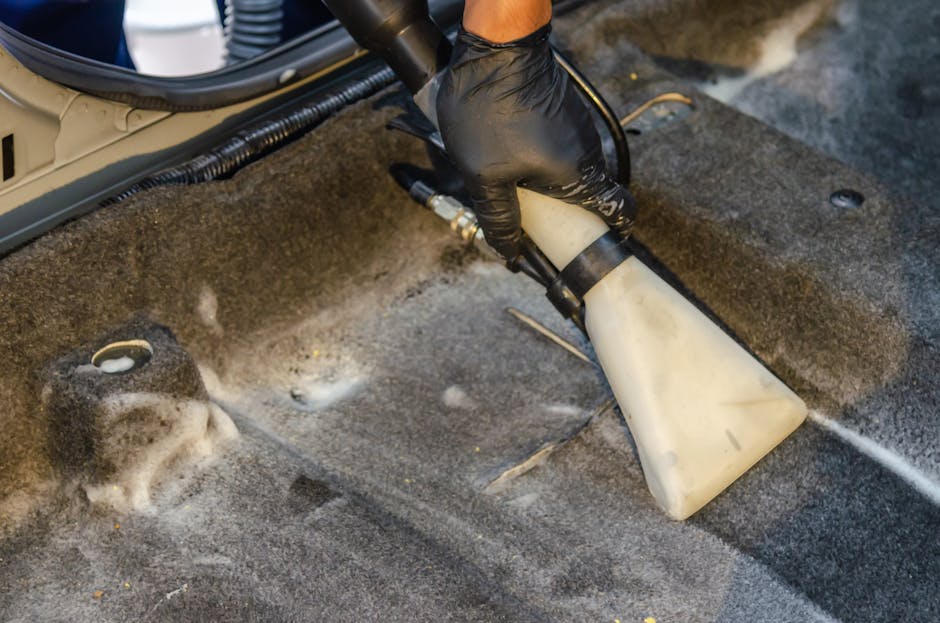 A professional cleaner dressed in a white protective suit, gloves, and boot covers is performing deep cleaning on a plush, beige carpet in a well-lit, modern living room. The cleaner is using a steam 
