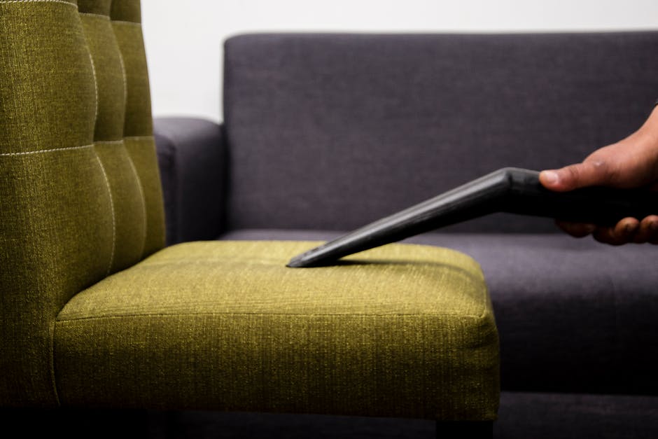 A person wearing gloves is using a handheld electric scrubber to deep clean the dark fabric upholstery of a sofa in a well-lit living room. The sofa has a modern design with a straight backrest and wi